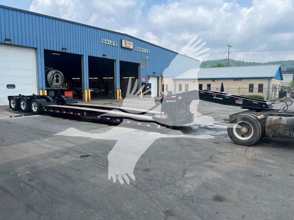 XL Specialized XL110HDGS lowboy trailer in a yard, attached to a semi-truck tractor, with a blue service building in the background.