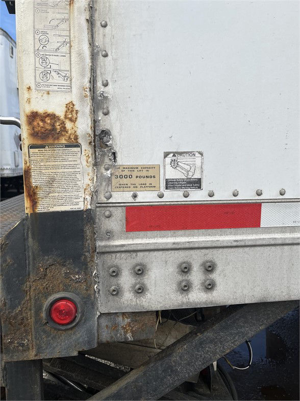 Rear corner of a metal box trailer with riveted panels, warning labels, and rust around a lower edge. A small label indicates capacity and a caution sign is visible.