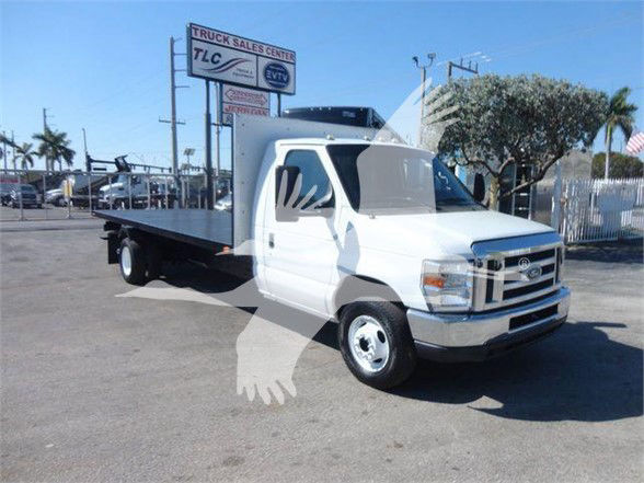 Flatbed tow truck in white with Ford emblem, long flatbed, cab-front view, dealership lot in background.