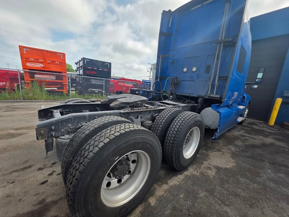 Truck with blue cab showing rear triple-axle chassis with dual rear wheels and fifth-wheel area. Visible components include frame rails, suspension, and fuel tanks.