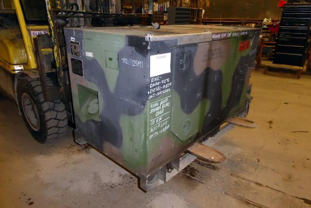 Truck-mounted generator unit with camo paint; various labels and handwritten notes on the side, including serial stickers and warning text.