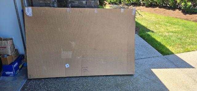 Large cardboard box on driveway outside with shadows and greenery in background.