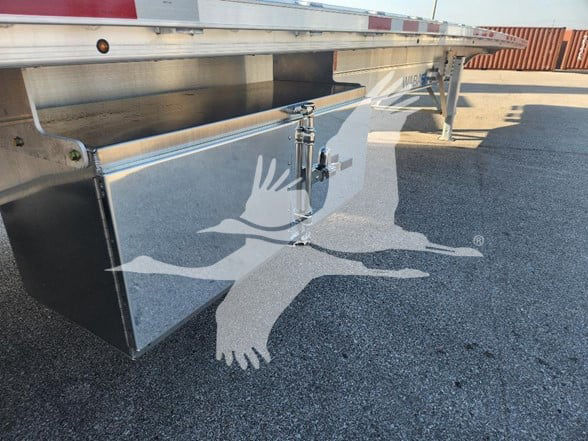 Trailer underside showing a chrome toolbox or storage box under a flatbed, with support legs and a reflective surface; watermark of a flying bird/logo overlaid.