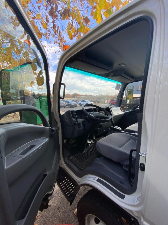 Isuzu NRR cab interior view from open driver door showing steering wheel, dashboard, and grey seats; exterior white cab and step, with autumn leaves in background.
