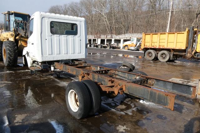 White cab of a 2007 Hino 145 chassis with exposed rusty frame and rear dual wheels, in a yard with other trucks.