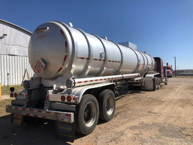 Tank trailer for crude oil transport with cylindrical stainless tank and rear loading equipment; red cab in background; hazard placard on rear left; long wheelbase with triple-axle configuration.