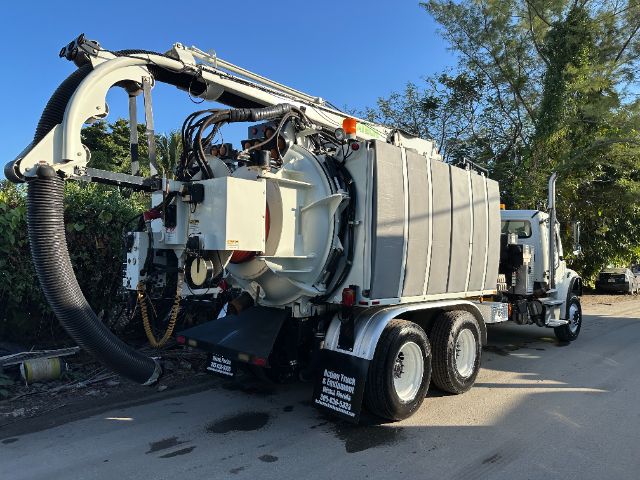 Freightliner M2 vacuum truck with large cylindrical tank and vacuum hose attachment on rear, multi-axle configuration, side mirrors, and blue sky.