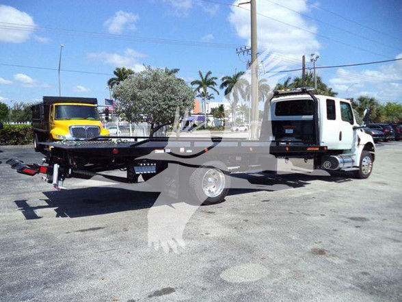 Tow truck flatbed with white cab and black bed, International MV chassis, early staging yard, power lines in background.
