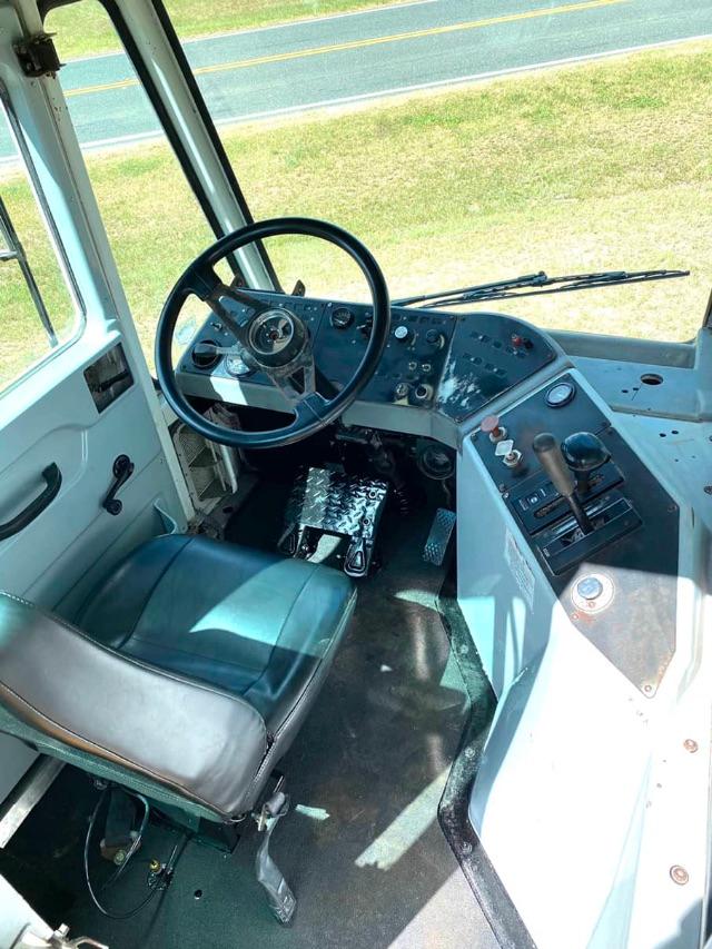 Bus cockpit showing steering wheel, dashboard with various gauges and controls, driver seat, and right-side console in a light blue interior.