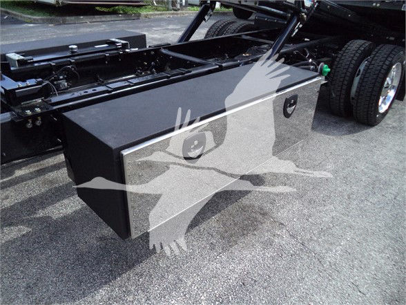 Truck chassis with a large rectangular metal toolbox mounted between rear wheels; side handles visible; black toolbox with reflective surface.