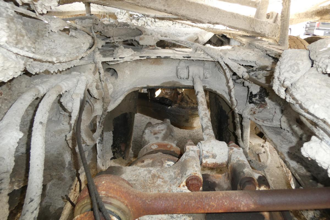 Tractor-compound view of a damaged Mack mixer chassis showing rusted drive shaft and framing with visible hydraulic lines.
