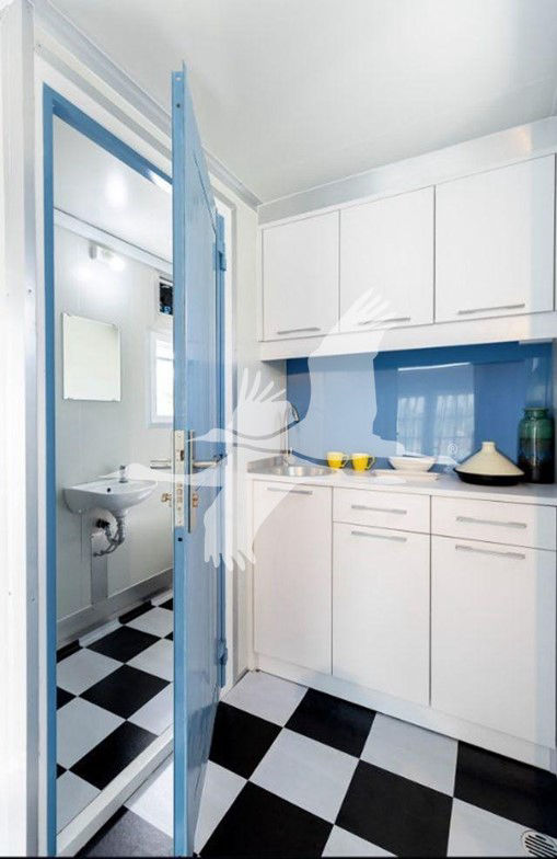 Interior of a trailer kitchen with white cabinets, blue backsplash, and checkered black-and-white floor. Open doorway to a small washroom.