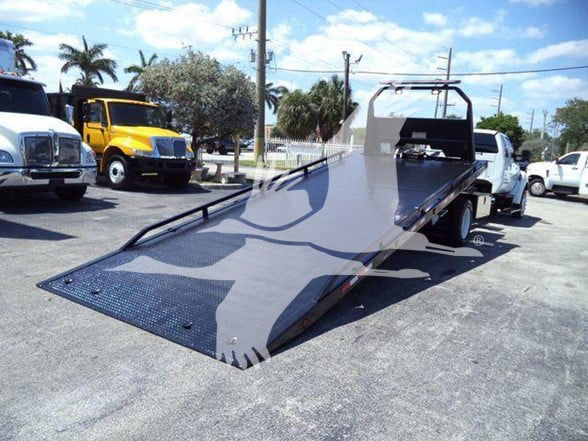 Black flatbed tow truck with a long inclined ramp in a dealership lot; other trucks visible in background.