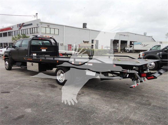 2023 International CV tow truck with flatbed, black dual rear wheels, truck cab in the background, industrial building and other vehicles.