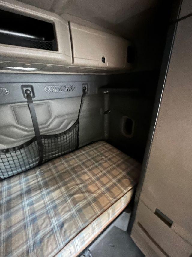 Cabin bunk area of a 2015 Freightliner Cascadia truck sleeper. Visible features include a plaid sleeper mattress, overhead storage shelf, windowless panel, and inlet straps.