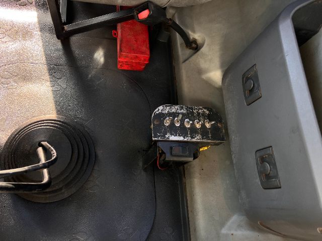 Truck interior view showing a red fuse/relay block, a black metallic dashboard panel with multiple small screws, and a gray plastic storage bin.