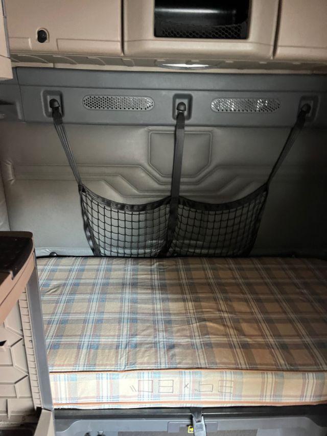 Cab interior of a Freightliner Cascadia sleeper showing a bed with plaid mattress, back wall with storage net and ventilation holes.