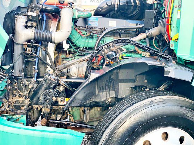 Volvo VNL 670 engine bay with exposed routing, hoses, and components; front wheel and fender visible; various cables and green engine cover amid servicing area.