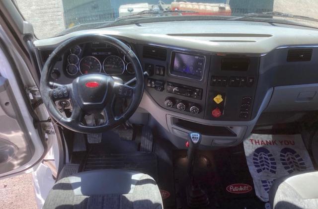Peterbilt truck interior showcasing steering wheel, instrument cluster, dashboard, gear shifter, and central touchscreen; visible floor mats and pedals.