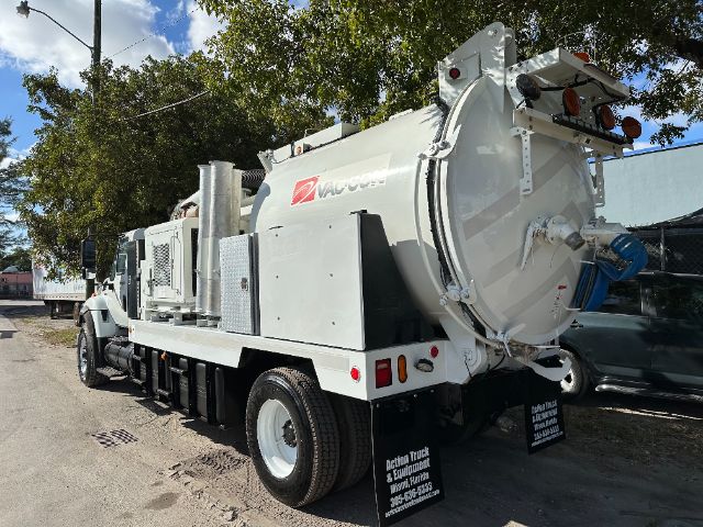 Rear view of a white vacuum truck with a large circular vacuum tank mounted on a long chassis. The tank shows a logo and text 'VAC-...'; rear equipment includes hoses, controls, and reflectors. Side panel with access doors and a diamond-pla