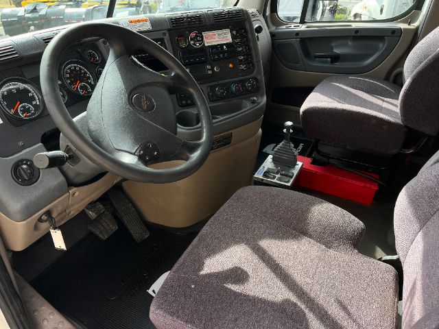Cab interior of a Freightliner Cascadia truck showing steering wheel, dashboard, gear shifter, pedals, and driver seat.