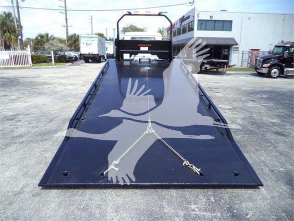 Flatbed tow truck bed with hydraulic ramp and chains, rear view showing rear bumper and surrounding lot.