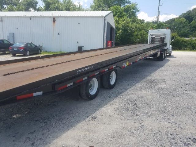 Flatbed trailer with long wooden deck and dual rear axles, semi-truck cab at far end, in a gravel lot.