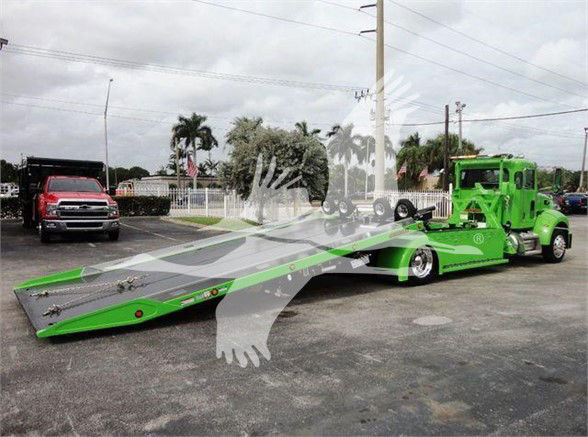Green Peterbilt tow truck on a car hauler trailer with low deck and extended ramp in a parking lot.