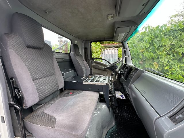 Isuzu NPR-HD cab interior showing driver seat, passenger seat, and center control console with switches. Dashboard and steering wheel visible.