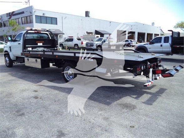 Flatbed tow truck in an outdoor lot with white cab and long flatbed, viewed from rear-right side.
