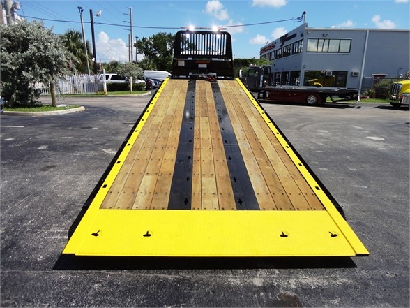 Tow truck flatbed with wooden planks and steel strips, yellow painted frame, headboard at far end, parking lot and building in background.