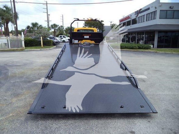 Flatbed tow truck ramp with a yellow loader in the background; turquoise building on right; logo silhouette of a bird across the ramp.