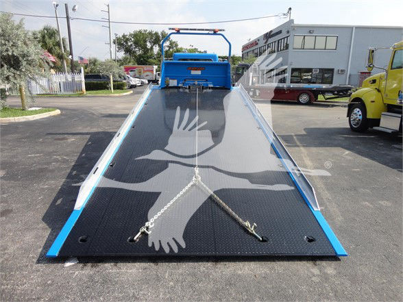 Tow truck flatbed with black diamond plate surface, blue frame, rear ramp, and safety chains stretched across. No legible labels visible on surface.