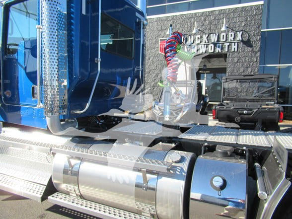 Kenworth W900L truck cab side with chrome steps and fuel tanks, blue paint, and dealership building in background.
