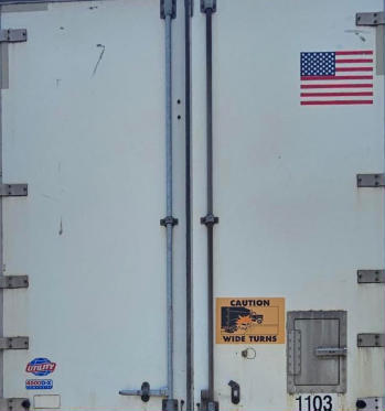 Rear of a white utility trailer with double doors, American flag sticker, CAUTION wide turns sign, serial/stock numbers 1103, 2014 Utility 4000 D-X Composite listing.
