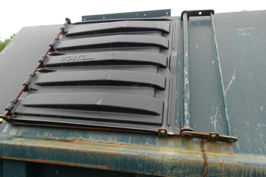 Wastequip dumpster lid with black hinge and latch on blue metal container, showing chain and rusted edge.