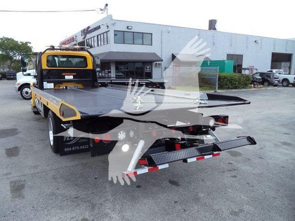 Tow truck with flatbed in rear, International DURASTAR 4300 style; visible markings include a yellow cab with black accents, board deck, and rear ramp. text on side may include contact numbers and company branding but is not fully legible.