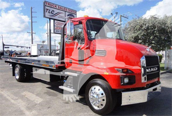 Red Mack MD6 tow truck with flatbed, chrome wheels, and dealership sign in background; visible text includes 'MACK' on grille, 'TLC' sign, and general site scenery.