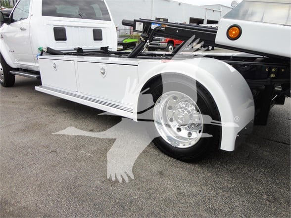 Ram 5500 Tow truck front side view showing white body panels, chrome wheel, and utility compartments with a mid-configuration hoist and flatbed on a dealership lot.