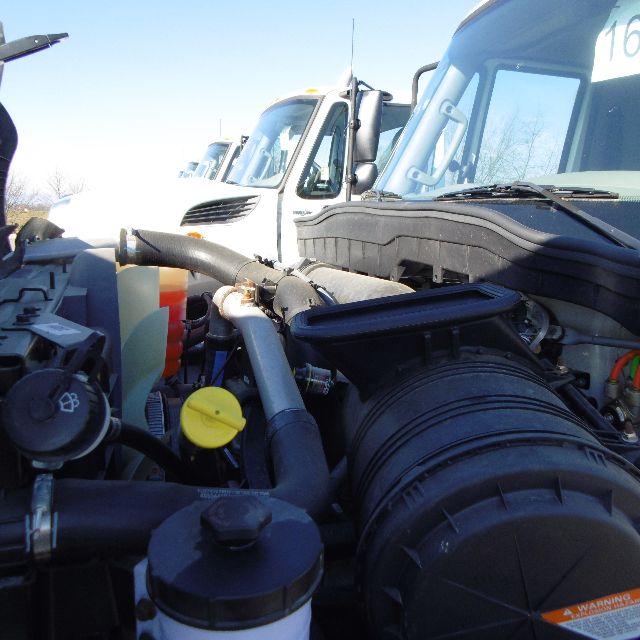 Engine bay of a 2016 International 7400 SBA 6X4 truck showing hoses, reservoir caps, and air intake with other trucks in background.