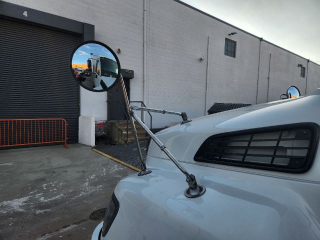 Truck hood with circular side mirrors and convex hood-mounted mirrors, front fender, and passenger-side window visible in industrial lot.
