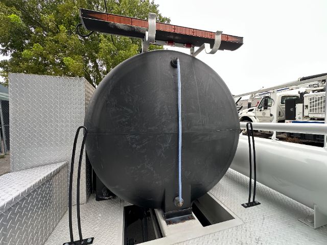 Flatbed truck with a large circular black tank mounted on the deck. A blue strap runs vertically across the tank. Diamond plate surface and a red light bar above. Background shows trees and other trucks.