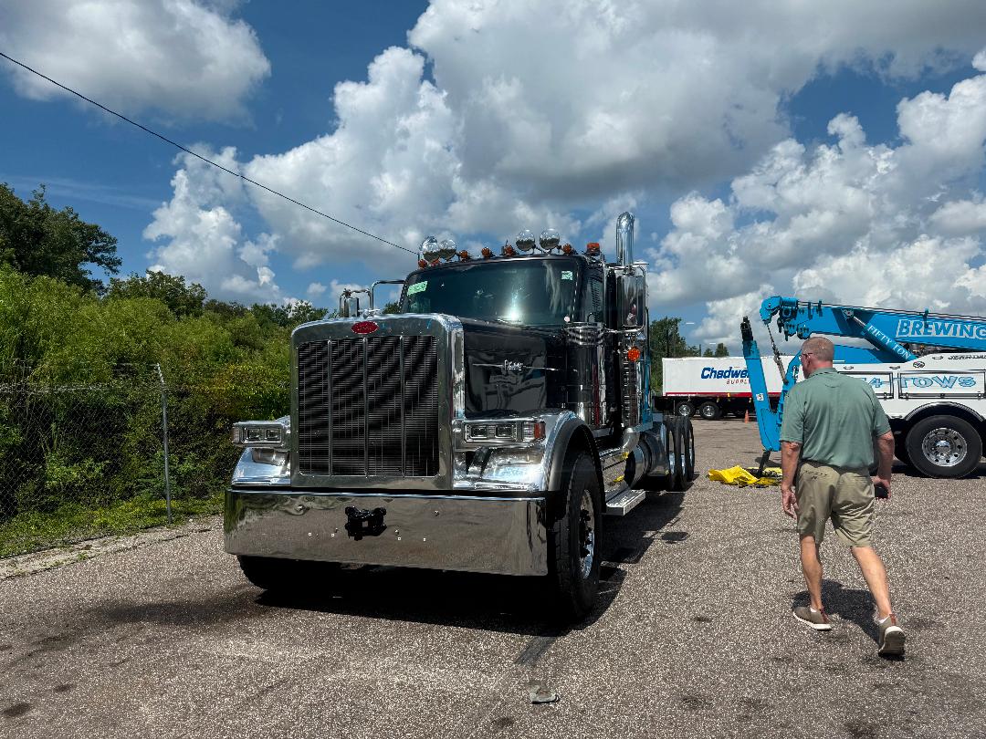 Peterbilt 589 heavy haul truck in a parking lot with chrome front grille, large bumper, and dual rear axles. A man in green shirt and shorts walks nearby. Clear blue sky with clouds.