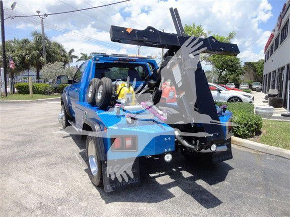 Tow truck with a blue body, rear-mounted hydraulic arm and winch. Visible labels and equipment on the back including spare tires and a yellow gas can, with a dealership watermark over the image.