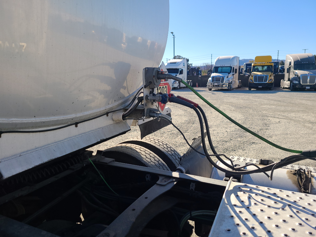 Semi trailer connection with jockey wheels and hoses at the rear of a Beall semi-trailer. Electrical and air hoses attached to a towing hitch with a red warning plate visible. Other trucks in the background.