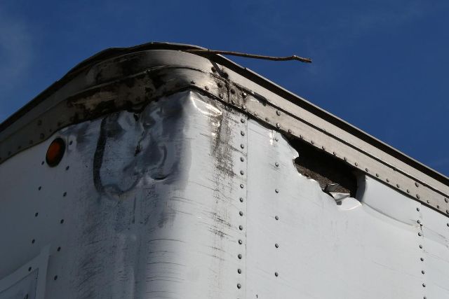 Image shows the corner of a metal trailer with riveted seams, dented edge, and chipped paint, top edge with a black weather seal.