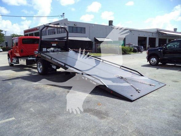 Tow truck with a flatbed trailer in a dealership lot, red truck in background, with a white building and cloudy blue sky.