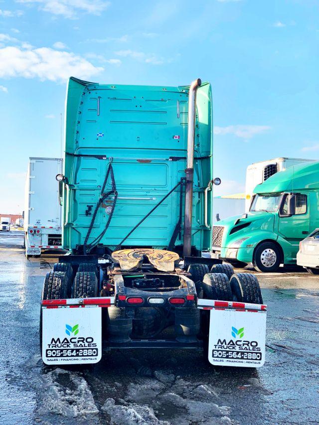 Rear view of a teal Volvo VNL 670 semi-truck tractor with fifth wheel visible, parked in a lot. Mud flaps show Macro Truck Sales contact number.
