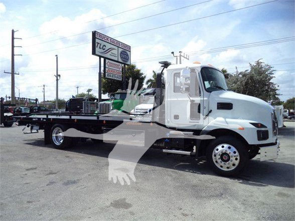 2024 Mack MD6 tow truck with flatbed, side view, white cab and wheeled rear, dealership lot in background.