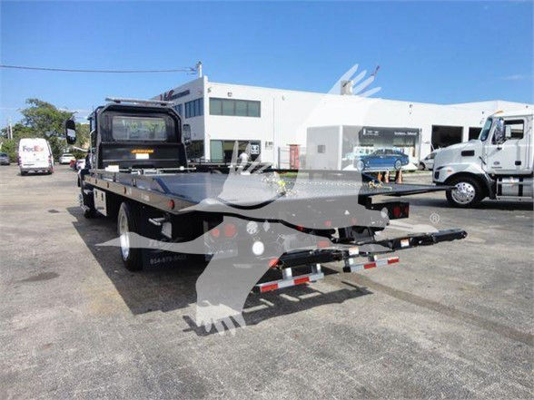 Flatbed tow truck with gooseneck hitch in a dealership lot, Mack MD6 platform visible, white cab in background, loading ramps at rear.